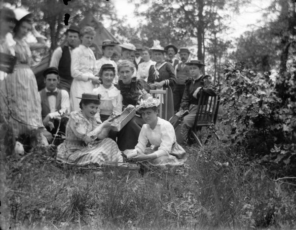 A large group of women gathered outdoors, possibly cutting potatoes. In the background is a tent.