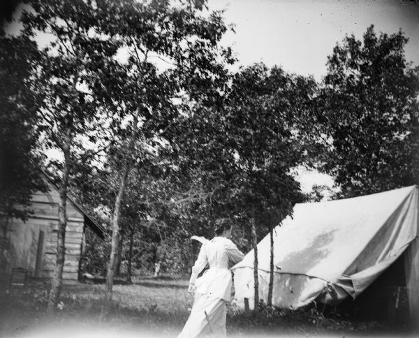 A woman walks toward a tent that is erected in a small grove of trees.