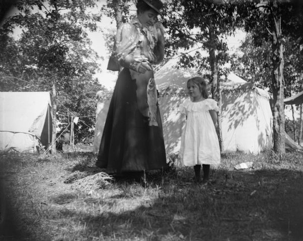 A woman displays a fish, probably a trout, by a string as a young girl looks on. Additionally, several large tents are erected in the background of the photograph.