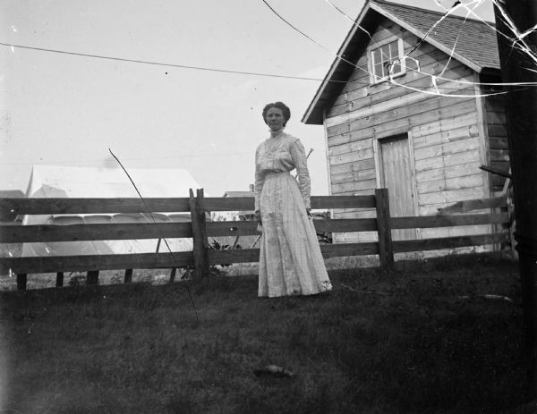 A woman stands near a large, white tent that is erected in a yard. There is a building on the right.