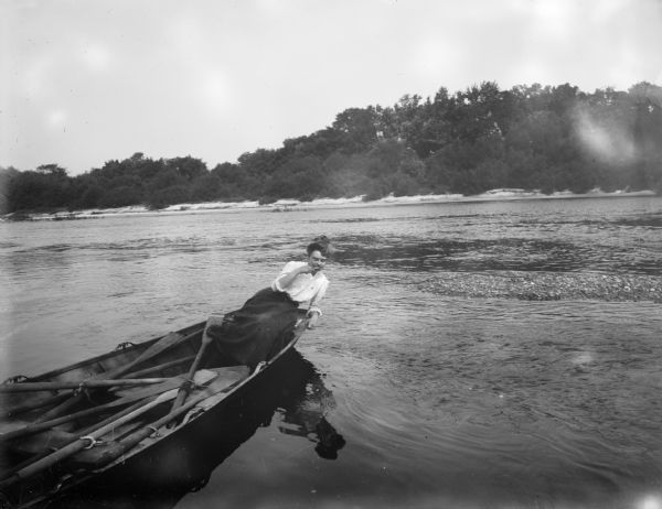 A woman sits and leans over the side of a wooden rowboat on a river. The far shoreline can be seen in the background.
