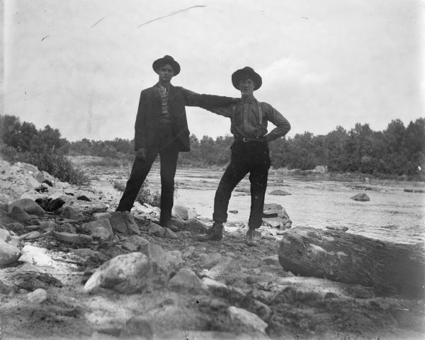 Two men stand with a hand on each other's shoulder on the bank of a river.