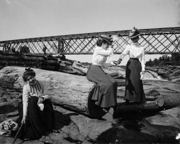 Three women playfully pose among logs near a railroad bridge that passes over a dam. On the bridge, a standing man is visible.
