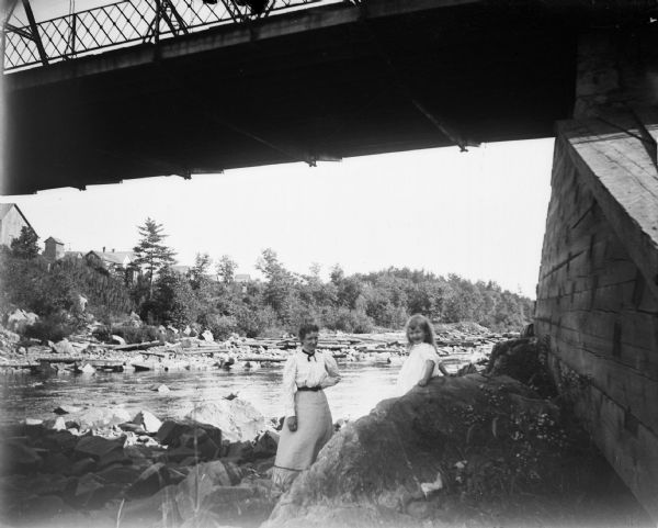 A woman and a girl, who sits on a large rock, pose underneath a bridge.