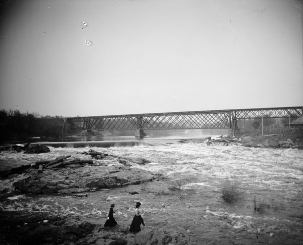 Two women walk the shoreline of a river. In the background, a dam and railroad bridge are visible.