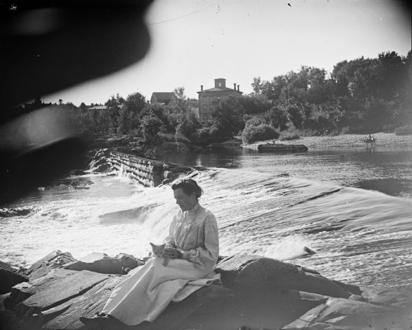 A woman sits with a kitten in her lap near a dam. This dam is probably the log dam on the Black River. In the background, the boarding house for Spaulding workers is visible.