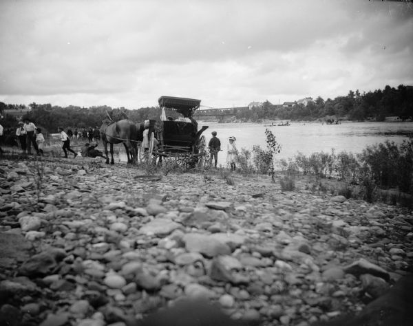 A group of people gather along the shore of a river with a horse-drawn vehicle. In the distance is a railroad bridge.