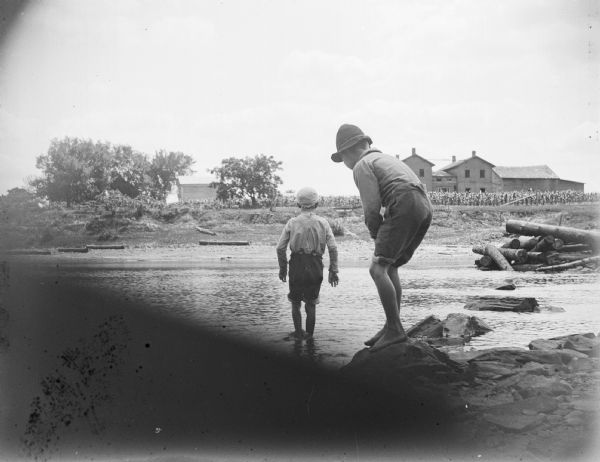 Two young boys wade across a river among logs, probably across the Black River from the Brockway building.