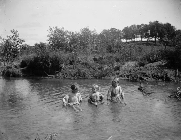Three small girls wearing dresses sit in a stream near a shoreline.