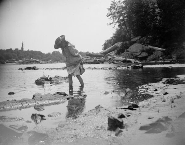 A young girl wades through a river and lifts the bottom of her dress, preventing it from getting wet.