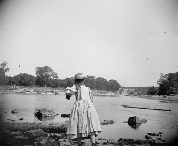 A young girl is standing at the edge of a shoreline looking down river, perhaps having just thrown a stone.