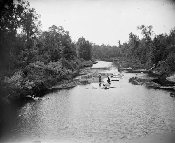 Taken from a distance, one woman stands on a sandbar while the other wades in the water at the narrow neck of a river.