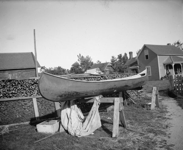 A canoe rests on sawhorses in front of a stack of firewood and frame house.