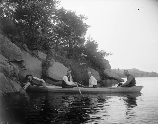 Four men sit in a boat near a rocky shore.