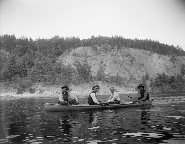 Four men sit in a rowboat on a river. In the background, a steep, rocky shoreline is visible.