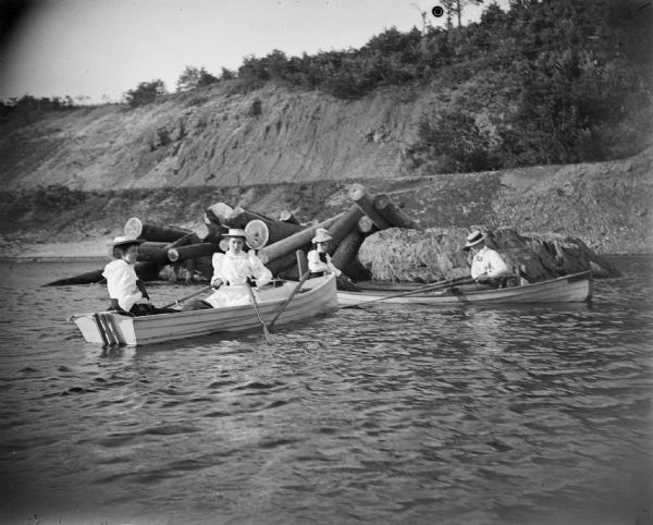 Four women, two in each boat, row near a pile of logs along a shoreline.