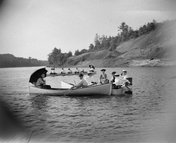 Three boats full of men and women row down a river. A steep shoreline is visible in the background.