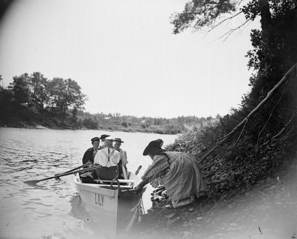 A girl pushes a rowboat named "Lily" away from the shore that has two women and two men seated inside.