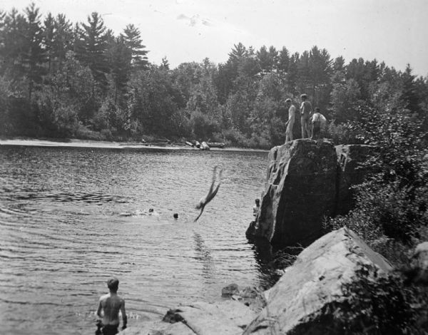 Elevated view of a boy diving into the river from a large rock, while others are awaiting their turn to jump. Several boys are already swimming in the water, possibly the Black River.
