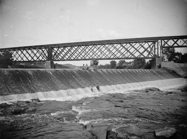 Several boys stand and sit on a concrete dam under a railroad bridge.	
