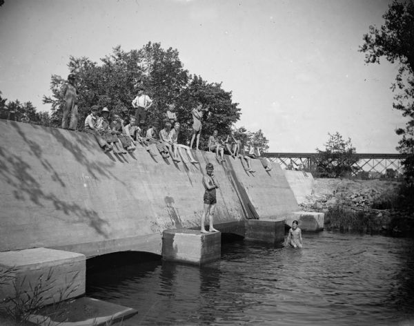 A large group of boys stand and sit on the bulkhead of a powerhouse before a flood. The boys could dive under one side of the structure and swim to the other side.