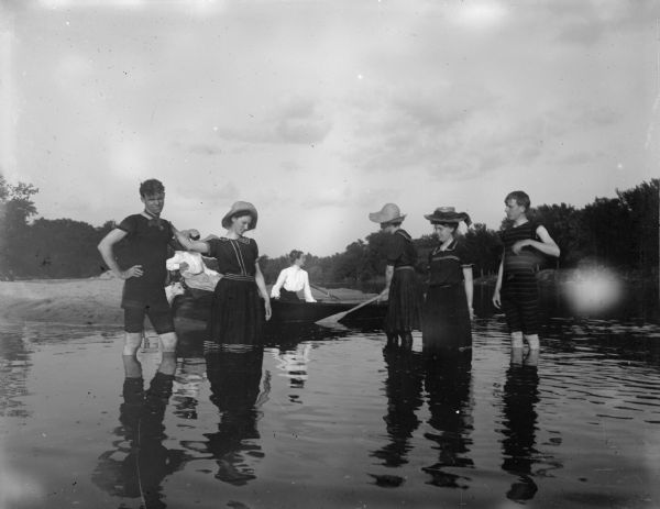 Three women and two men in swimming suits wade in the river, while two women sit in a rowboat on shore, possibly the Matt Olson farm campground.