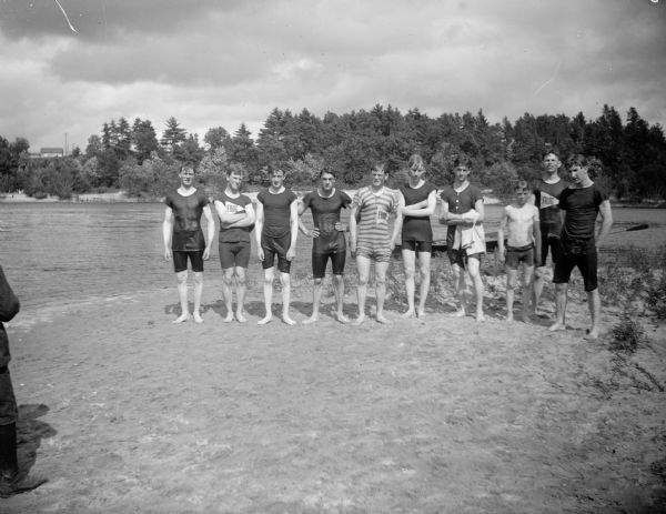 Ten young men in swimming suits pose in a line on a riverbank.