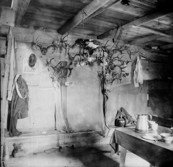 Interior view of a log cabin with antlers hanging on a wall. Items of clothing are hanging from the various antlers. Also, there is a table set with dishes.