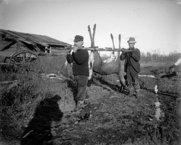 Two men carry a dead deer on a wooden rail. In the background is a log cabin.