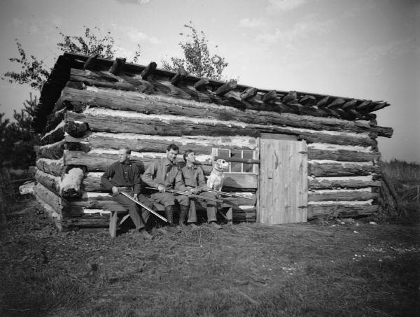 Three men with their guns and a dog sit on a bench outside of a log cabin. The man in the center is probably Virgil Taylor, who ran Rock Spring Bottling Works.