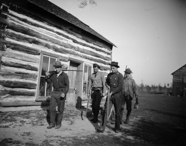 Four men with firearms walk outside a log cabin, one carrying a dead bird.