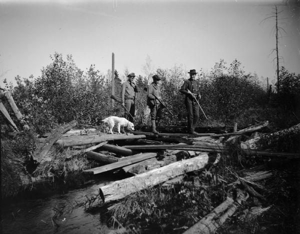Three men with firearms cross a stream with a dog following.