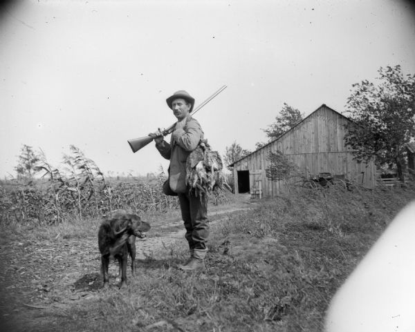 A hunter, probably John Sprester, stands in farmyard with his gun, dog and catch of prairie chicken. In the background, a barn is visible.