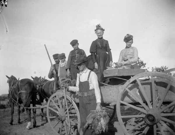 A hunter stands near a wagon pulled by two horses holding a catch of squirrels. Two women, two young boys, and another man are standing in the wagon behind him.