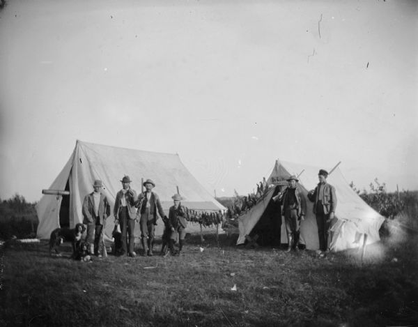 A group of five men and a boy pose with their hunting dogs and guns in front of a two-tent hunting encampment. A rail of fowl, possibly prairie chicken, hangs between the two tents.
