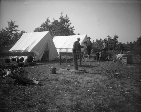 Four men pose in a two-tent encampment with a dog and various campground implements.