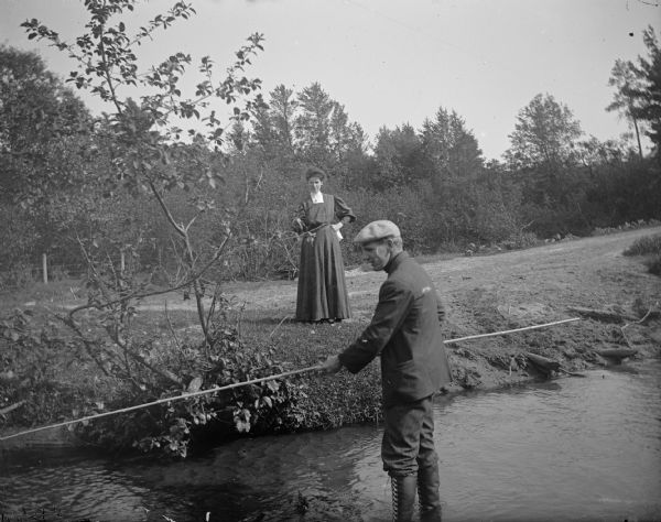 A woman stands on the shoreline nearby while a man wades in a stream to fish.
