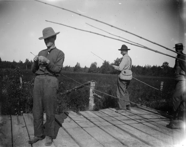 Three men bait the hooks of their cane fishing poles. They are standing on a wooden pier.