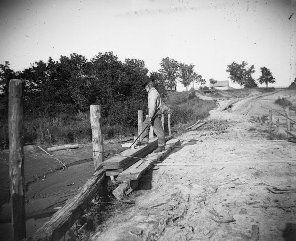 A man fishes from an earthen bridge. There is a house or barn in the background.