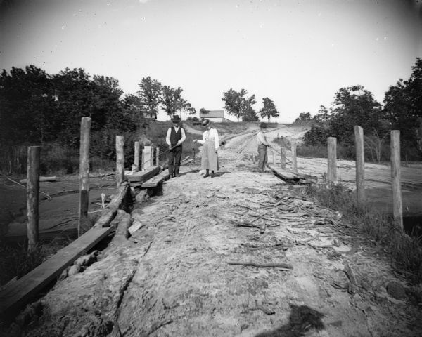 Two men fish from an earthen bridge while a woman stands holding a fish. There is a house or barn in the background.