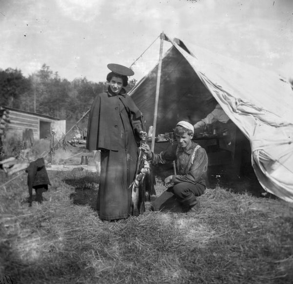 A young man and woman hold a string of fish outside a tent.