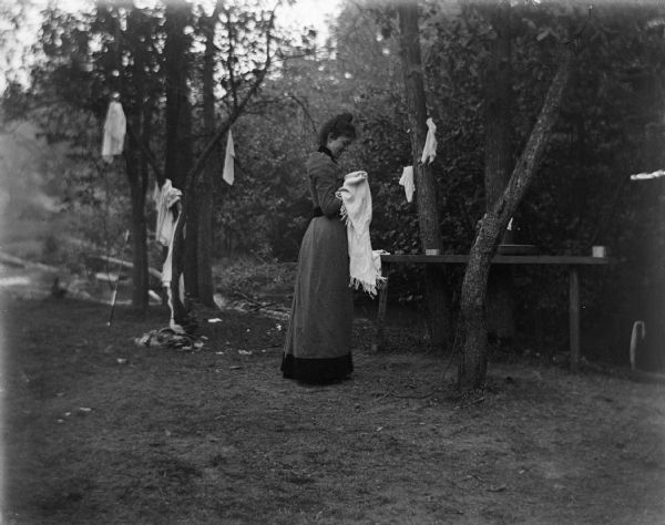 A woman stands with a towel near a campsite. Various other towels are hanging from the surrounding trees.