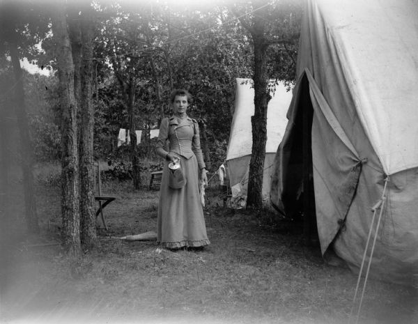 A woman stands near two tents at a campsite and holds a coffee pot.