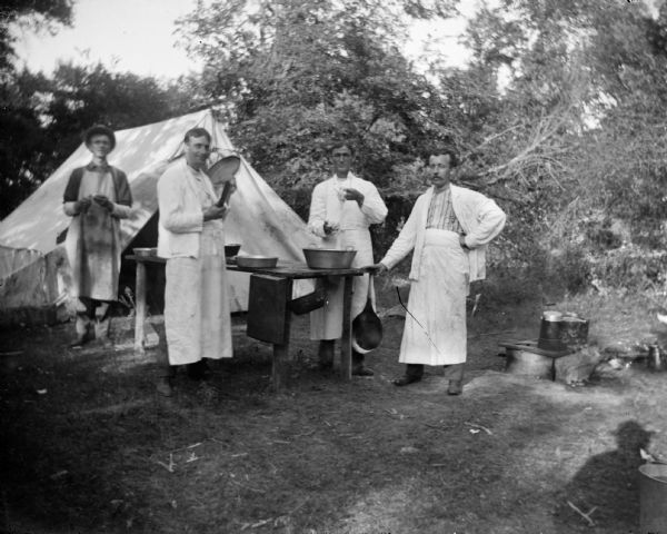 Four men wearing white aprons attend to kitchen duties outdoors at a campsite. There is a tent in the background.