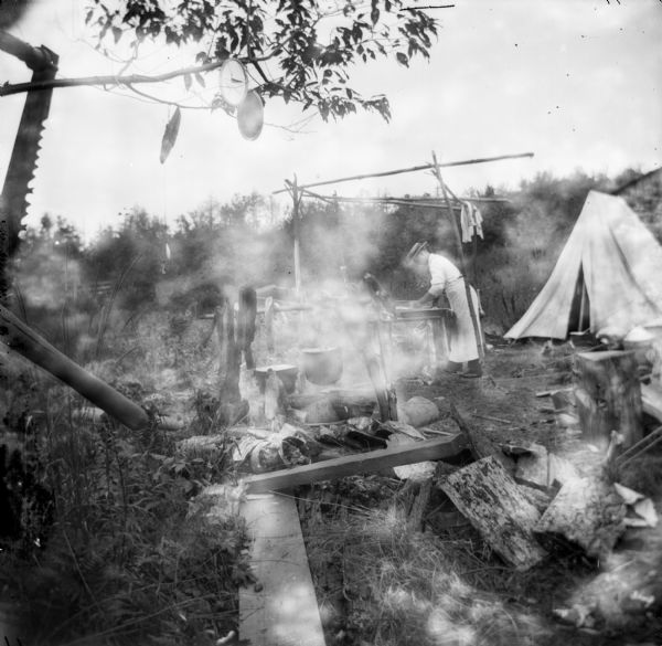 A man wearing an apron cooks over an open fire at a campsite near a tent. Smoke or sunlight obscures the image.
