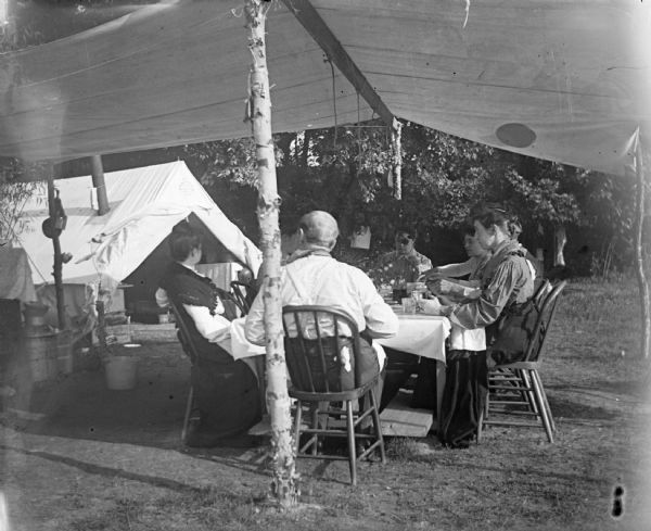 A group of men and women eat at a table underneath a tent awning.