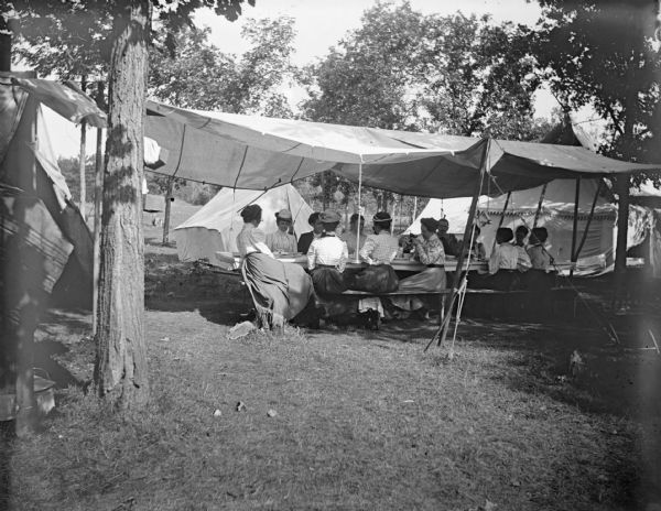 A large group of people sit outdoors at a long table underneath an awning. Probably at the Matt Olson Farm Campground, presently the site of Perry Creek Park.