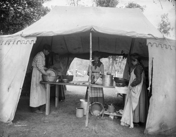 Two women and a man wash dishes in a tent.