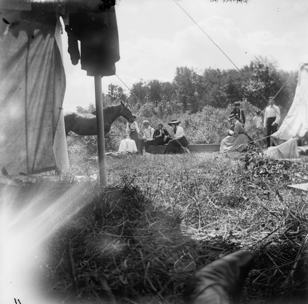 A group of men, women and children, as well as a horse, gather outside of two tents at a campsite. The photograph appears to have been taken from inside one of these tents.