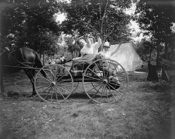 Three women wave as they sit on a wagon pulled by a horse.